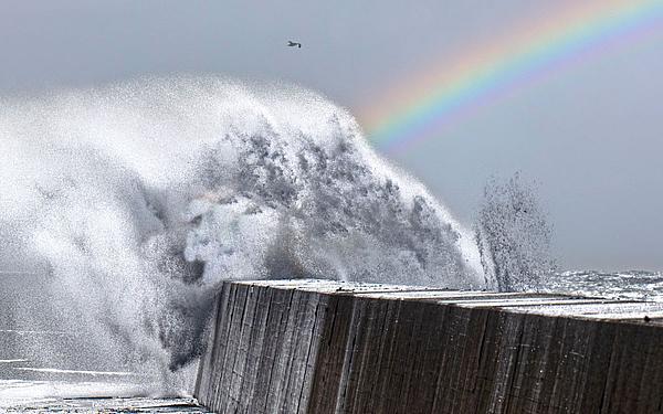 Eine dramatische Welle schlägt gegen eine Mole auf Helgoland, über ihr schwebt ein Regenbogen