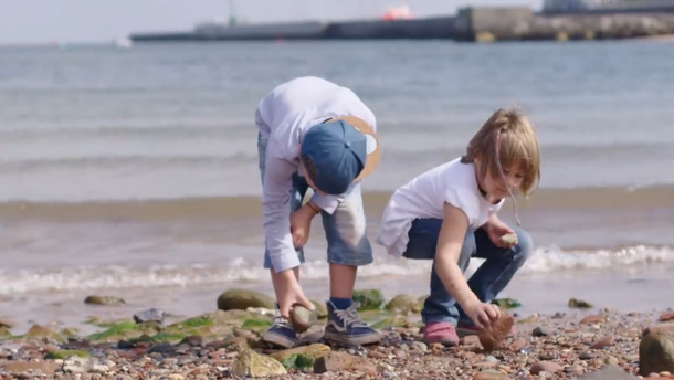 Zwei Kinder sammeln Steine am Strand, das Meer und eine Mole im Hintergrund.