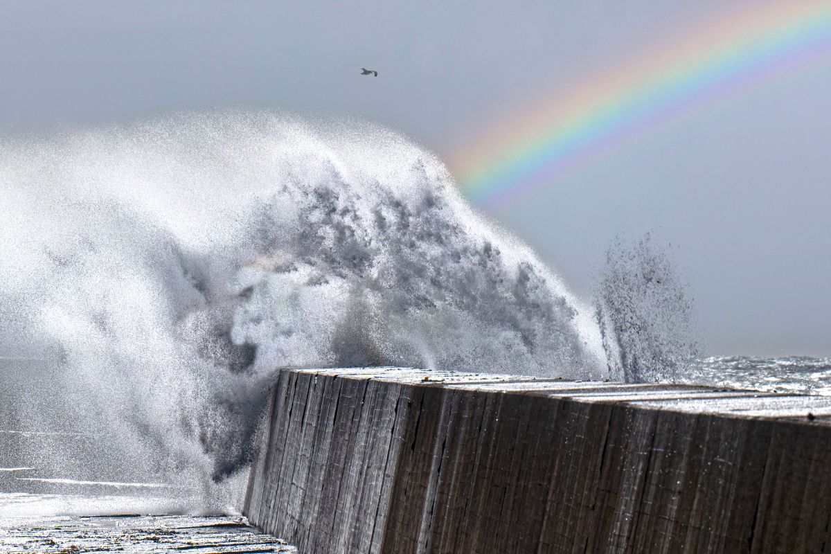 Eine dramatische Welle schlägt gegen eine Mole auf Helgoland, über ihr schwebt ein Regenbogen